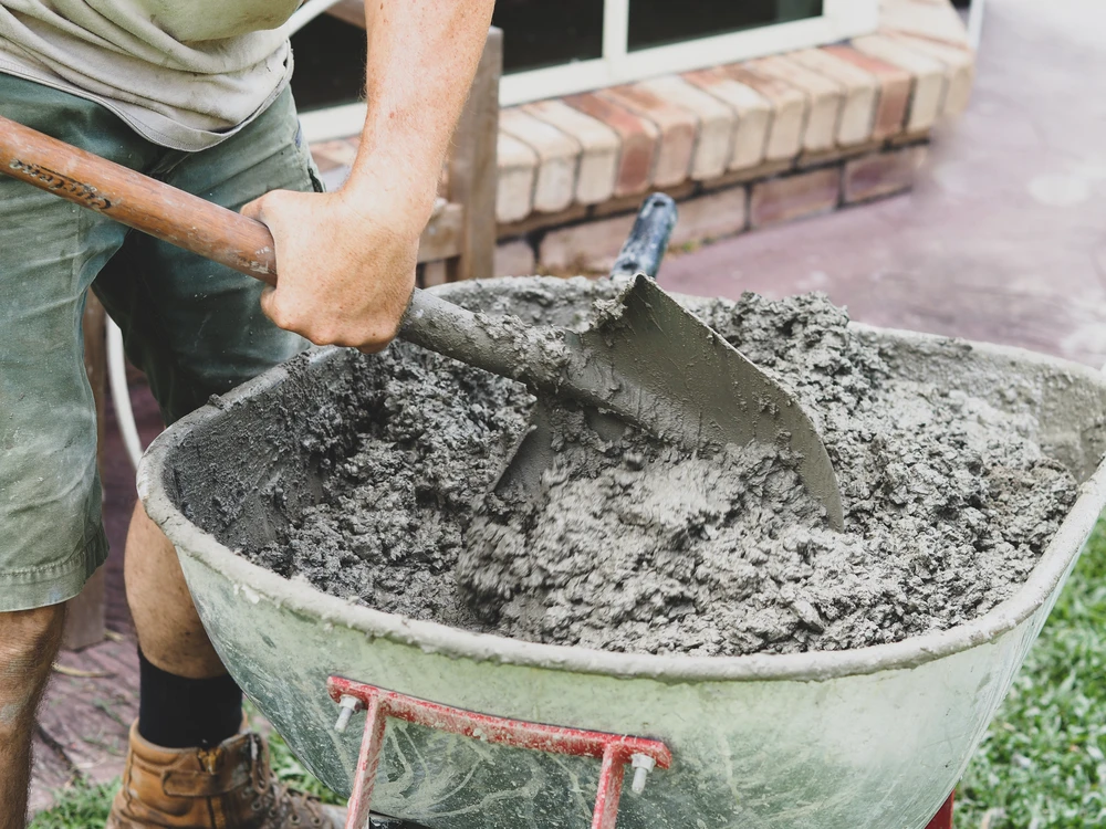 Worker mixing fresh concrete in a wheelbarrow using a shovel during a residential construction project.