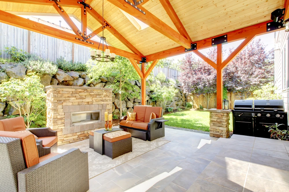 Covered outdoor living space with wood ceiling, stone fireplace, seating area, and grill in a landscaped Potomac MD backyard.