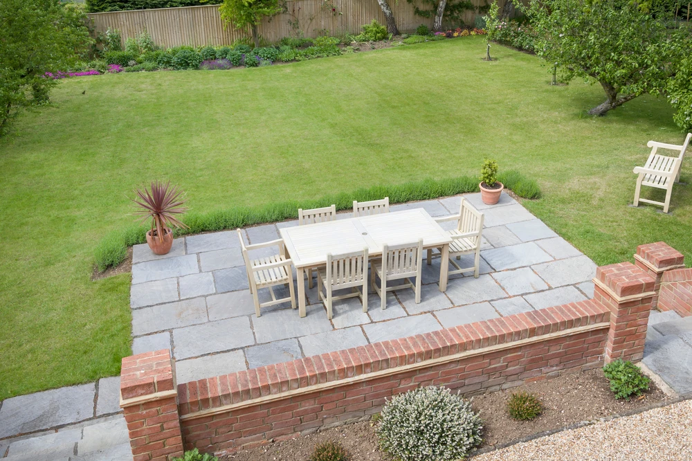Elevated stone patios with outdoor dining set overlooking a large green lawn and landscaped residential backyard.