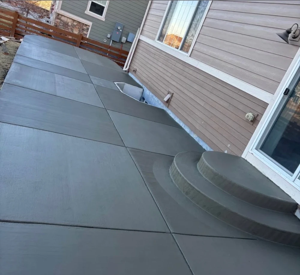 New concrete patio with curved steps follows the back of a gray house beside a wood privacy fence.