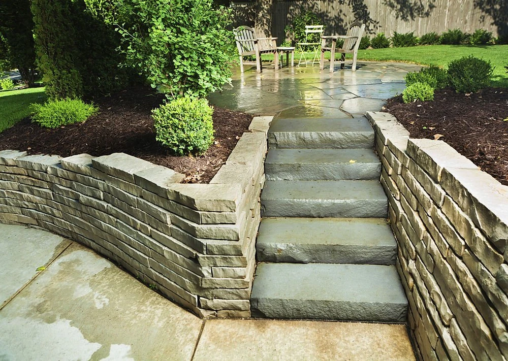 Stone steps and retaining walls lead to a refreshed backyard patio with chairs and trimmed shrubs.