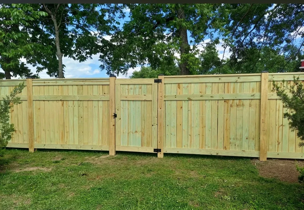 Tall wooden privacy fence with a double gate across a grassy yard under leafy trees.