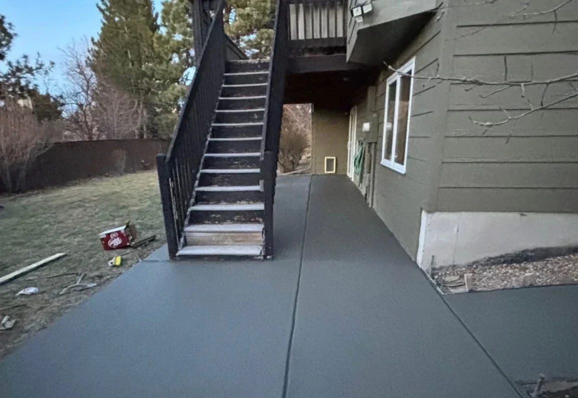 Concrete walkway under a black exterior staircase along a gray house with a large yard.